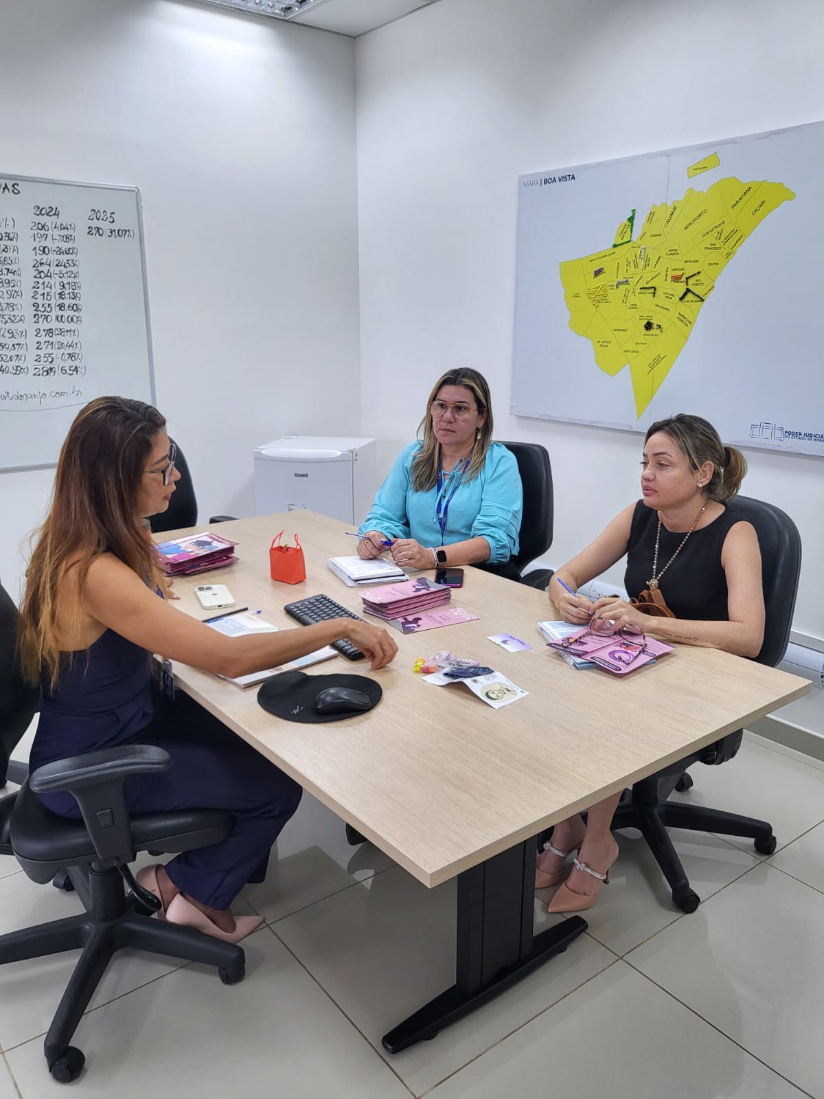 Imagem colorida mostra três mulheres sentadas a mesa. Na mesa tem algumas cartilhas Imagem colorida mostra três mulheres sentadas a mesa. Na mesa tem algumas cartilhas