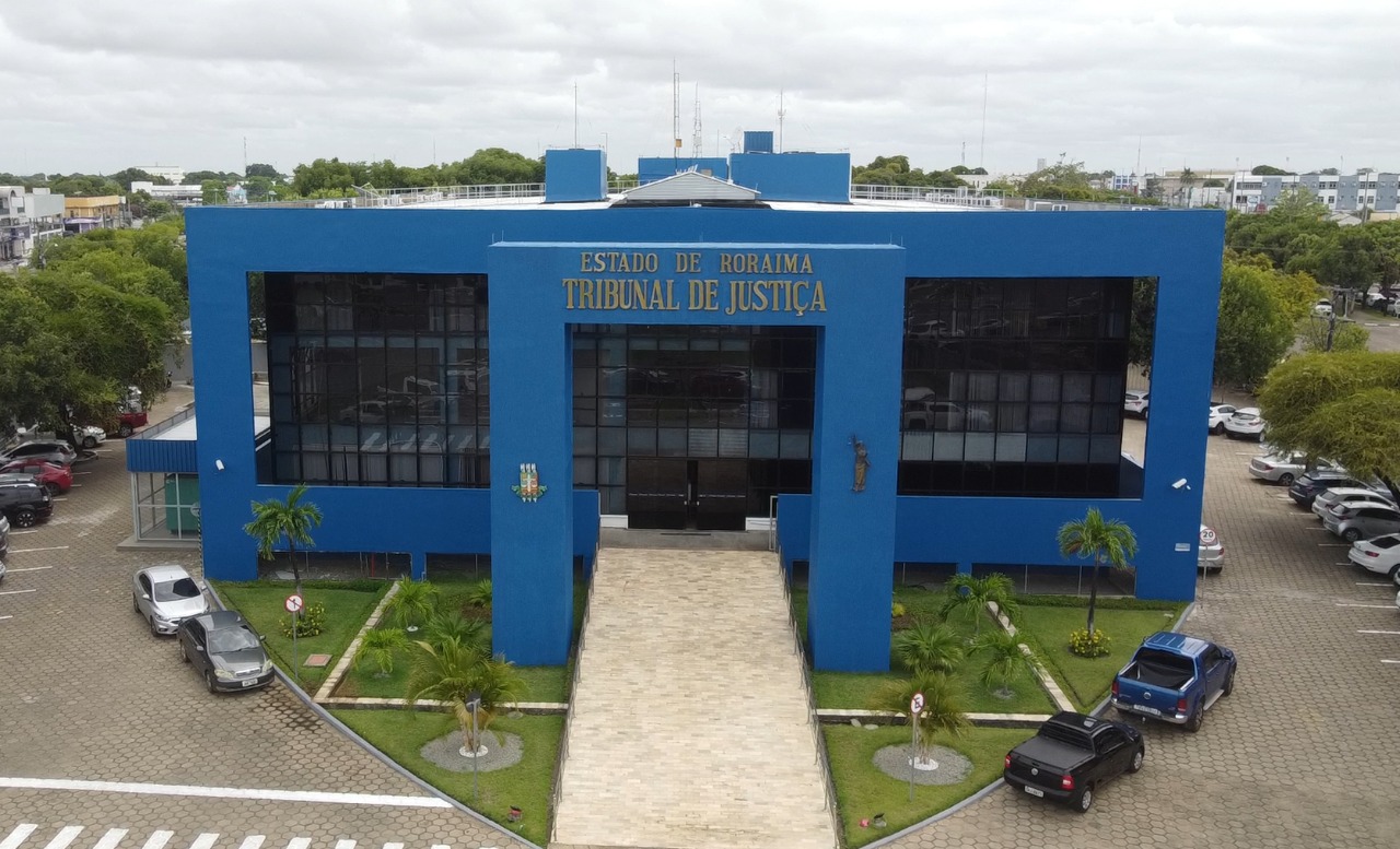  imagem aérea colorida  em formato retangular mostra o prédio do Palácio da Justiça e seu o estacionamento com diversos carros e uma pequena  área de vegetação ao redor do prédio. 