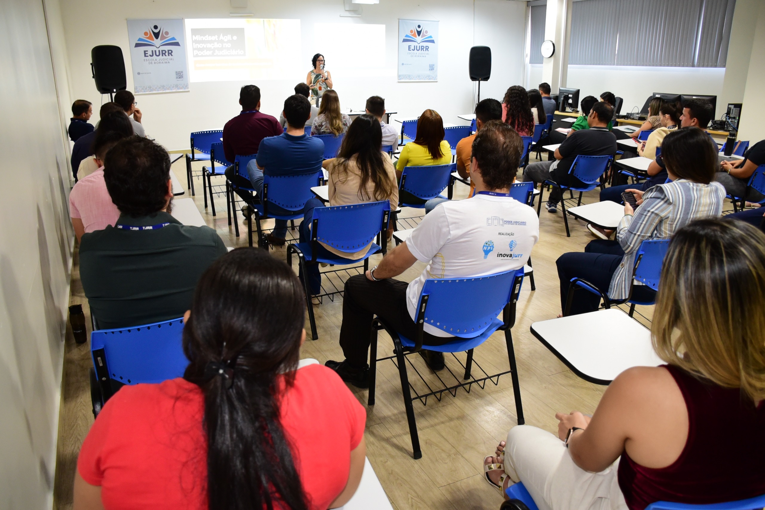 Foto colorida mostra um público de homens e mulheres sentados em cadeiras escolares, de costas para a câmera. Ao fundo está uma mulher em pé com o microfone na mão. Abaixo o texto “INOVAÇÃO – Programação com palestras, workshops e inaugurações reúnem mais de 400 participantes”.