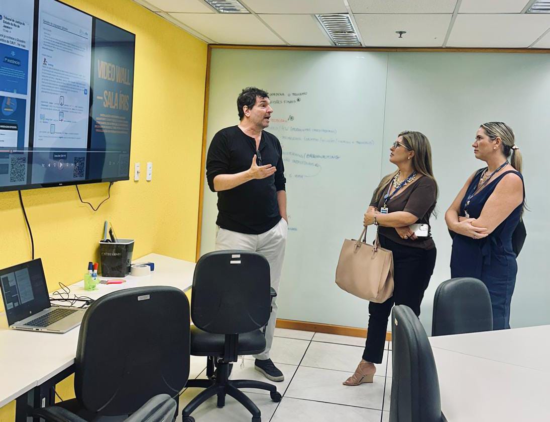 A imagem colorida mostra as servidoras Erika Horta e Cristine Helena Rodrigues, do Tribunal de Justiça de Roraima (TJRR)e mais um servidor do TJRJ conversando, durante uma visita técnica ao Tribunal de Justiça do Rio de Janeiro (TJRJ). Abaixo a frase “INTERCÂMBIO DE BOAS PRÁTICAS: Servidoras do TJRR realizam visita técnica ao TJRJ na área de Gestão por Processos e Qualidade.