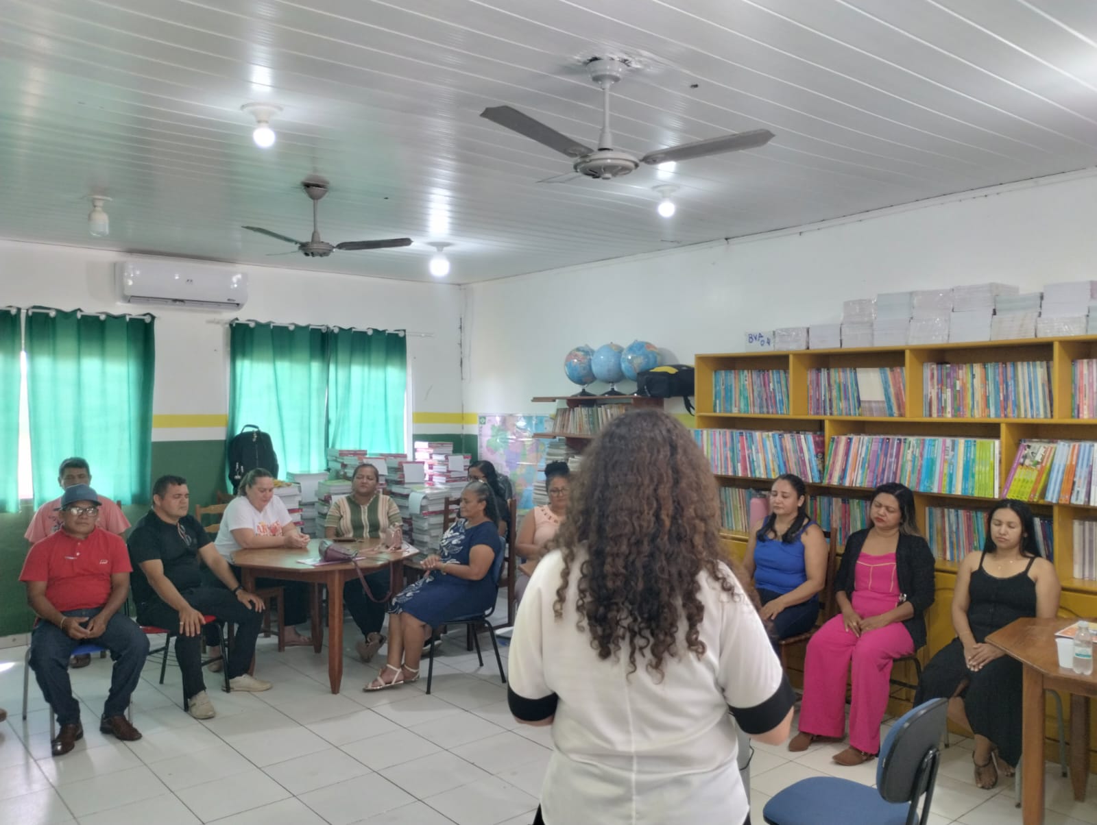 Foto colorida em um ambiente de sala de aula. No meio da foto há uma pessoa de cabelo cacheado virada de costas e ao fundo 11 pessoas sentadas em formato de meia-lua.