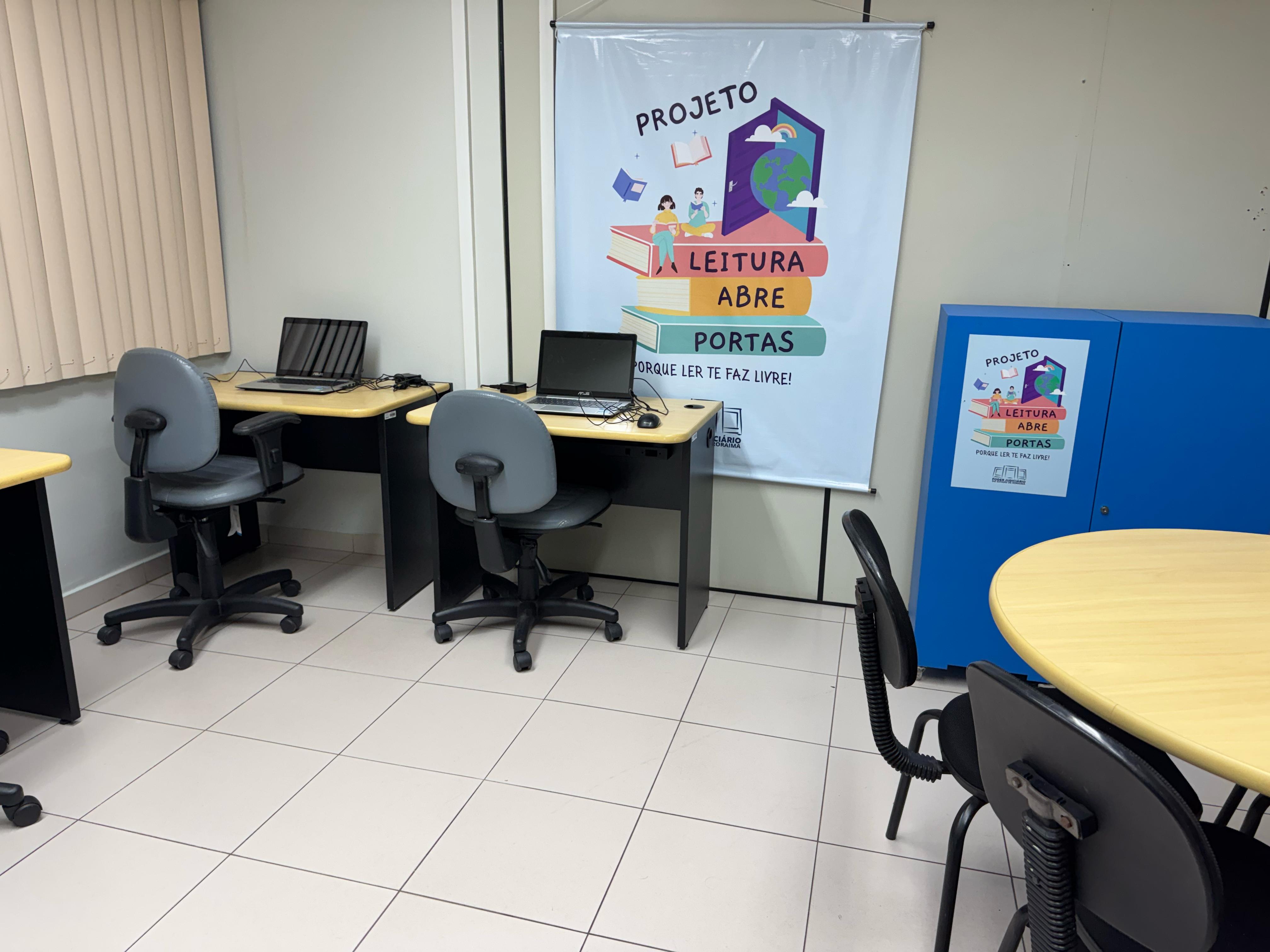 A imagem mostra uma sala com mesas, cadeiras giratórias e notebooks. Há um banner e um armário azul com o cartaz do projeto "Leitura Abre Portas". A imagem mostra uma sala com mesas, cadeiras giratórias e notebooks. Há um banner e um armário azul com o cartaz do projeto "Leitura Abre Portas".