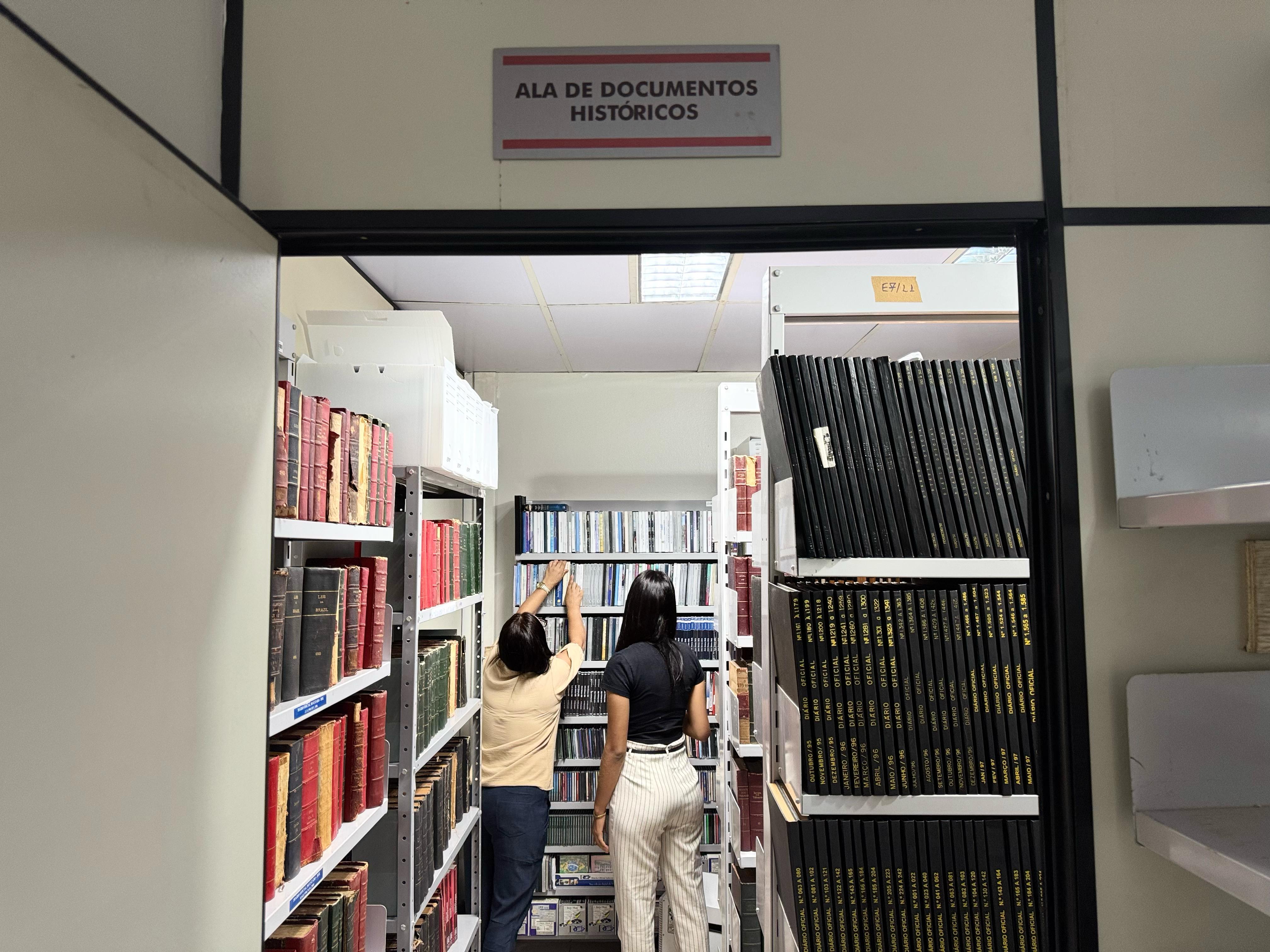 A imagem mostra a entrada da "Ala de Documentos Históricos", com estantes cheias de livros. Duas pessoas estão dentro do espaço, consultando materiais nos arquivos. A imagem mostra a entrada da "Ala de Documentos Históricos", com estantes cheias de livros. Duas pessoas estão dentro do espaço, consultando materiais nos arquivos.