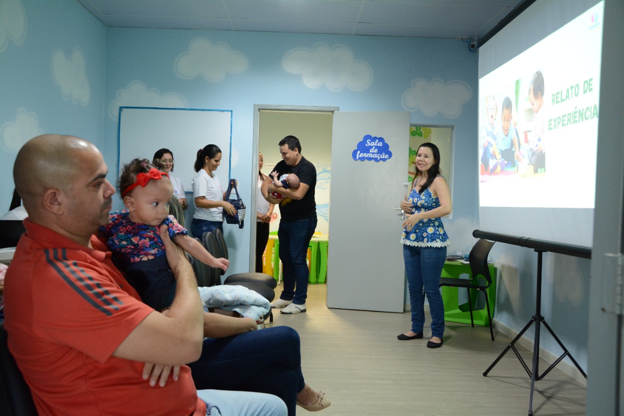 Imagem colorida mostra pessoas reunidas em sala durante palestra. Ao canto esquerdo da imagem um pai sentado segura sua filha bebê,  no lado direito a palestrante está em pé ao lado do slide no telão e no fundo as servidoras recebem um pai que está a porta em pé segurando seu filho.  