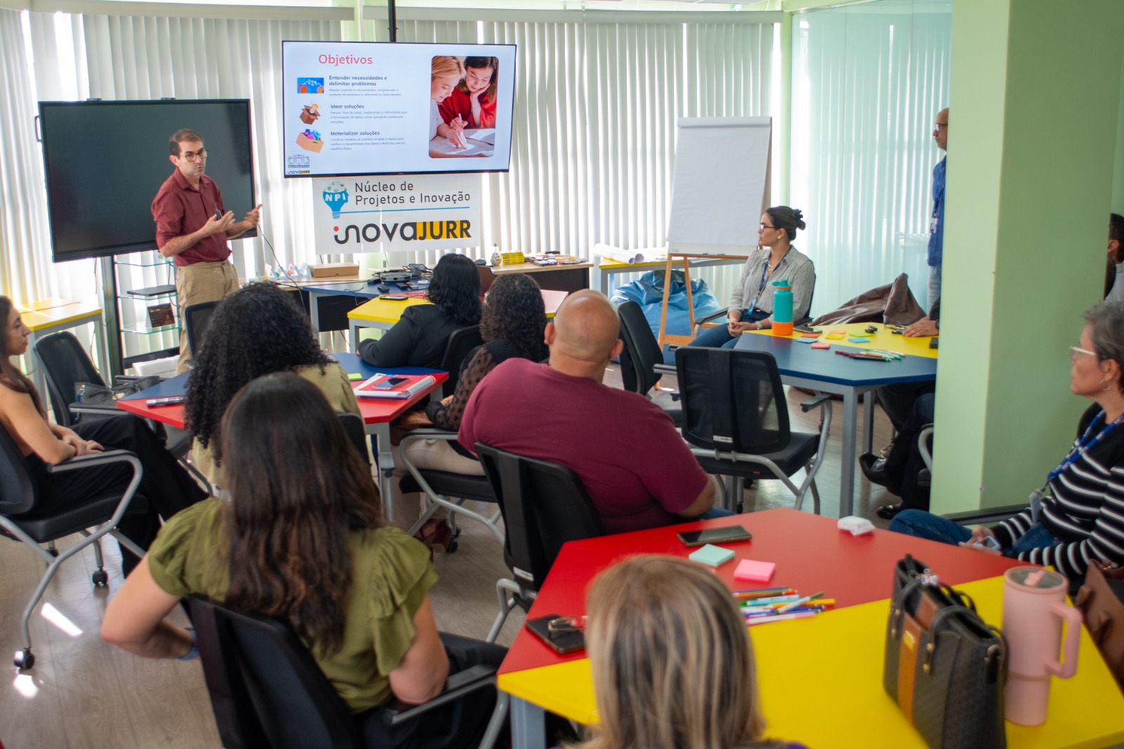 Foto colorida mostra uma sala de reunião com pessoas sentadas ao redor de mesas coloridas, assistindo a uma apresentação. Um homem em pé explica o conteúdo exibido em uma tela sobre objetivos de inovação. Foto colorida mostra uma sala de reunião com pessoas sentadas ao redor de mesas coloridas, assistindo a uma apresentação. Um homem em pé explica o conteúdo exibido em uma tela sobre objetivos de inovação.