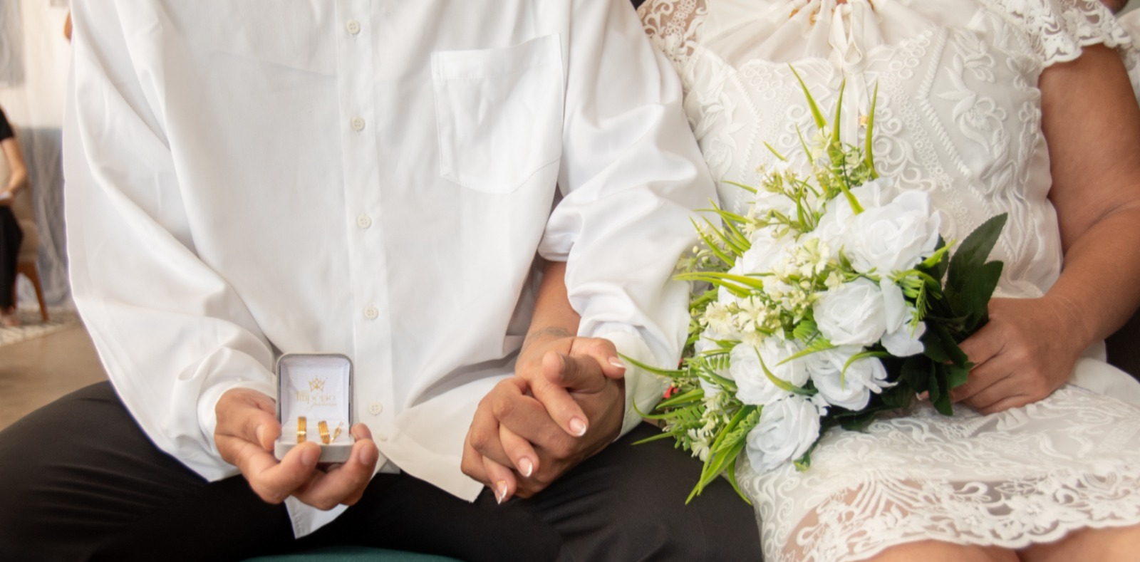 imagem em close-up de um casal sentado, com o foco da cintura para baixo. O homem (à esquerda) veste uma camisa branca de mangas longas e calça preta e segura na mão esquerda um estojo de alianças aberto. A mulher (à direita) veste um vestido de noiva branco, com detalhes de renda na saia, e segura um buquê de flores brancas com folhagens verdes. As mãos do casal estão entrelaçadas sobre o colo. imagem em close-up de um casal sentado, com o foco da cintura para baixo. O homem (à esquerda) veste uma camisa branca de mangas longas e calça preta e segura na mão esquerda um estojo de alianças aberto. A mulher (à direita) veste um vestido de noiva branco, com detalhes de renda na saia, e segura um buquê de flores brancas com folhagens verdes. As mãos do casal estão entrelaçadas sobre o colo.