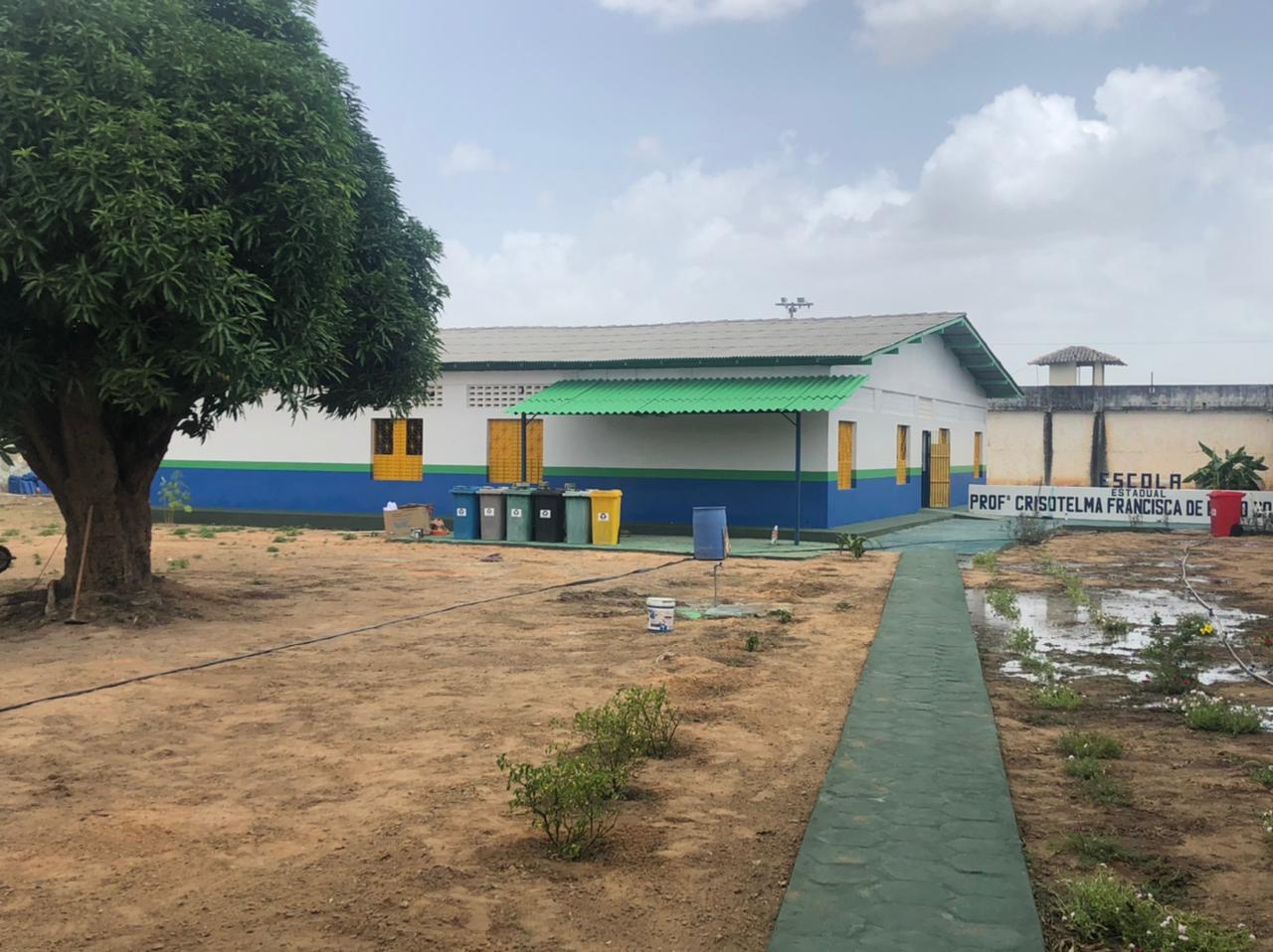 Foto da unidade de ensino dentro da penitenciária, ao lado dela uma árvore, uma calçada com algumas plantas dos lados e no fundo um muro.