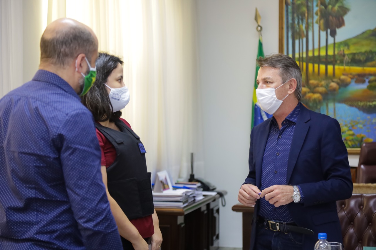 Foto da juíza titular da Vara de Execuções Penais, Joana Sarmento, e o governador Antônio Denarium de pé conversando, em uma sala. Ao fundo há uma mesa com papéis em cima, algumas cadeiras, uma bandeira do Brasil e um quadro na parede.