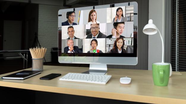 Imagem de uma mesa com computador, mostrando fotos de pessoas em reunião virtual, sobre a mesa há um recipiente com lápis, uma agenda e um celular do lado esquerdo, e no lado direito uma xícara e um abajur.