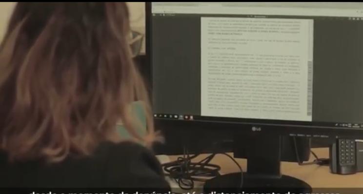 Foto de mulher de costas sentada em frente a um computador, lendo um texto que está na tela.