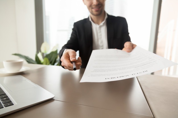 Imagem de um homem, entregando uma caneta e um papel, ele está atrás de uma mesa que têm uma xícara e um notebook em cima dela, ao fundo da foto uma janela grande e uma planta aparecem desfocadas.