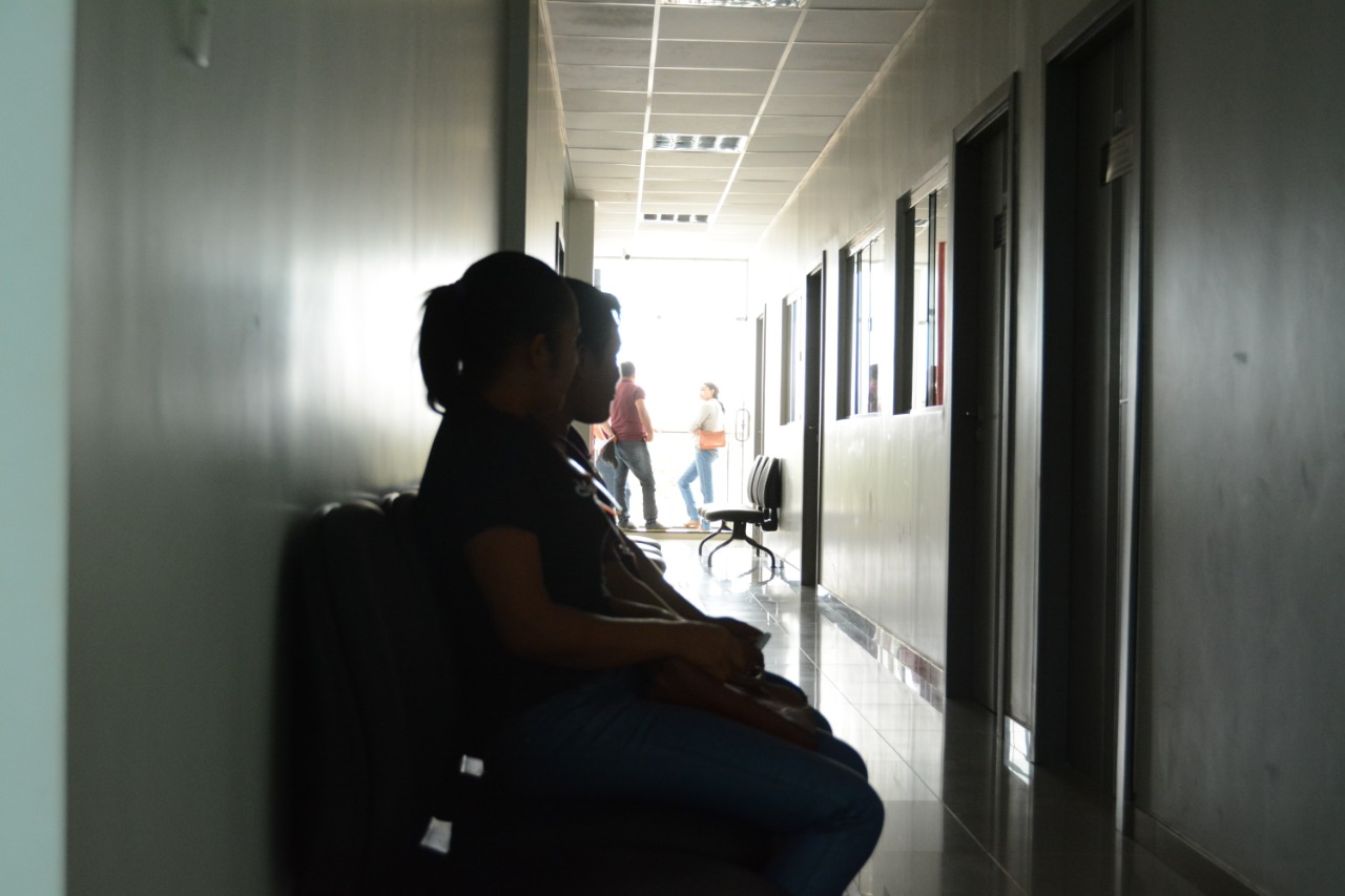 Foto de duas pessoas sentadas em cadeiras que estão em um corredor escuro, ao fundo do corredor há três pessoas de pé em frente a uma porta.