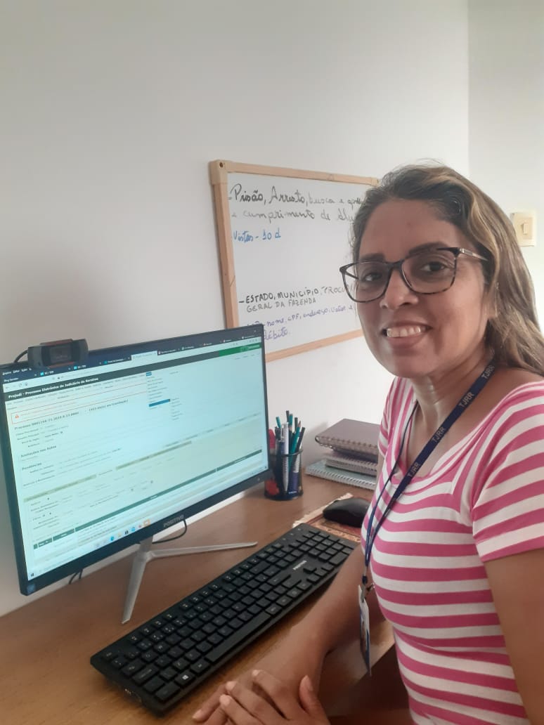 Foto da técnica judiciária Liliane Cristina Silva e Silva, sentada de frente á um computador que está sobre uma mesa que também têm canetas e livros em cima dela,no canto. na parede do fundo há um quadro branco. 