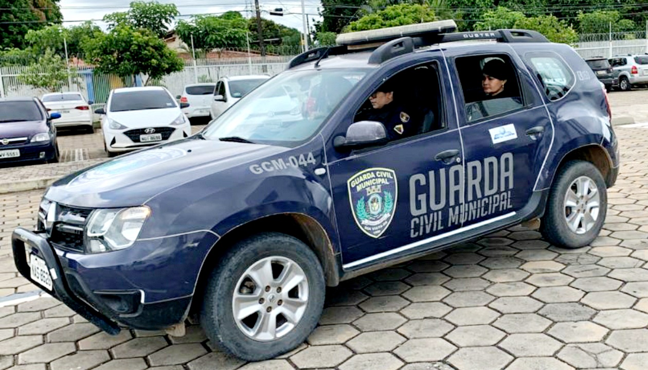 Foto de uma viatura da guarda civil municipal, com guardas dentro dela, e ao fundo um estacionamento de carros.