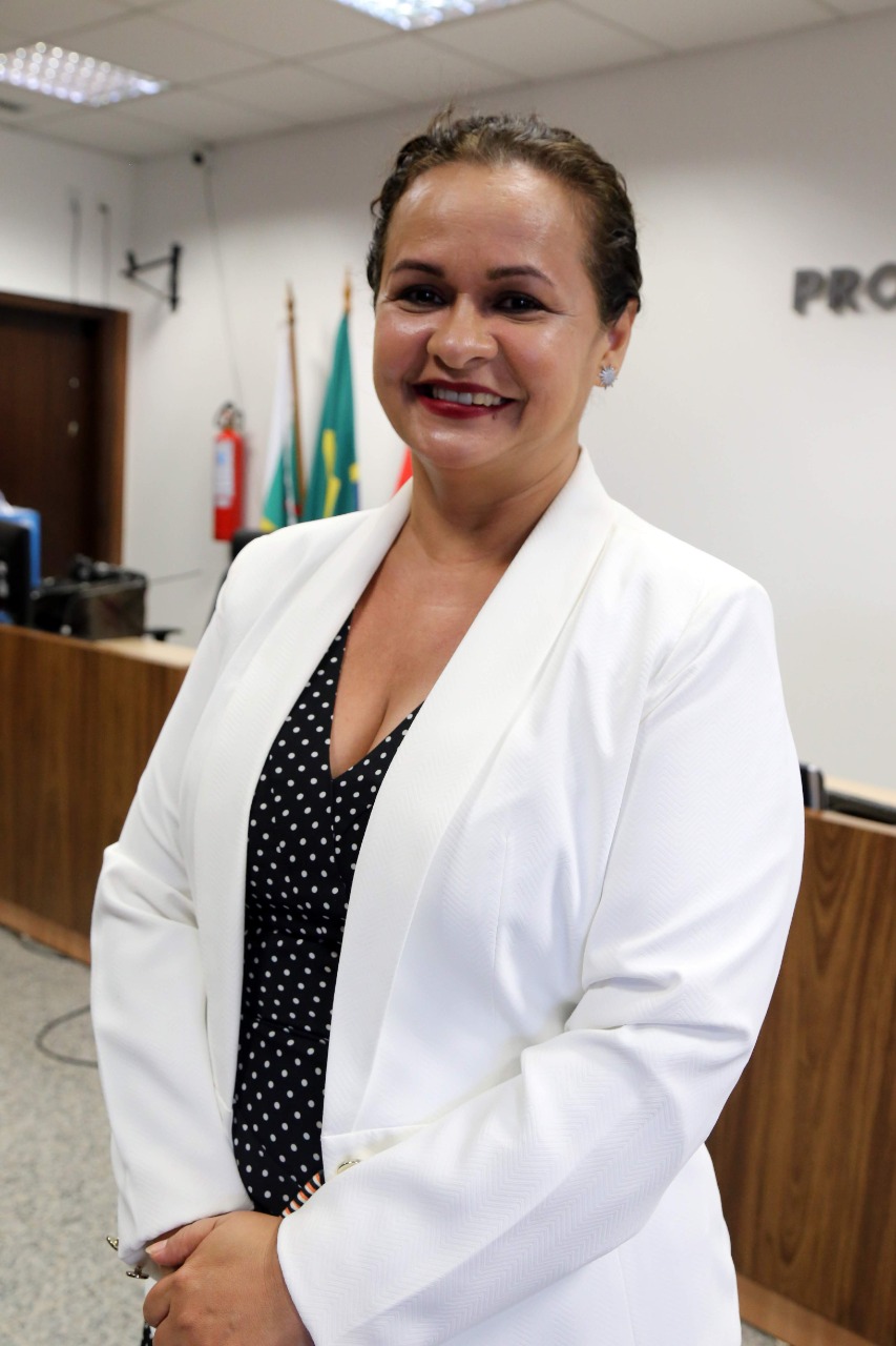 Foto da juíza Graciete Sotto Mayor Ribeiro de pé sorrindo, em frente à uma bancada, com bandeiras no canto da foto. 