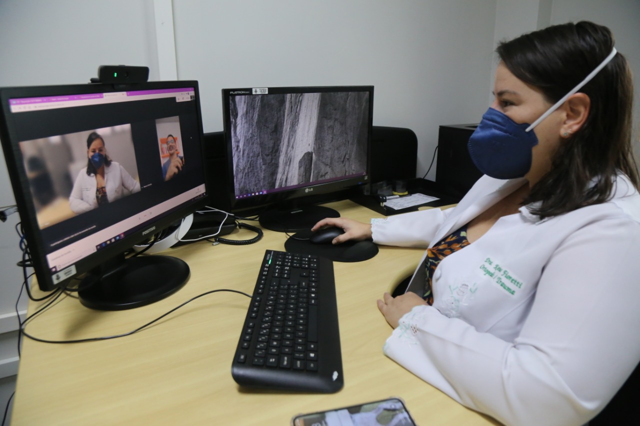 Chefe do Escritório de Saúde, Larissa Maravalha, sentada em cadeira vestida de jaleco e com máscara no rosto, segurando um mouse de computador que está sobre a mesa, que também têm um celular e duas telas de computador, em uma delas aparece a imagem dela em videochamada.