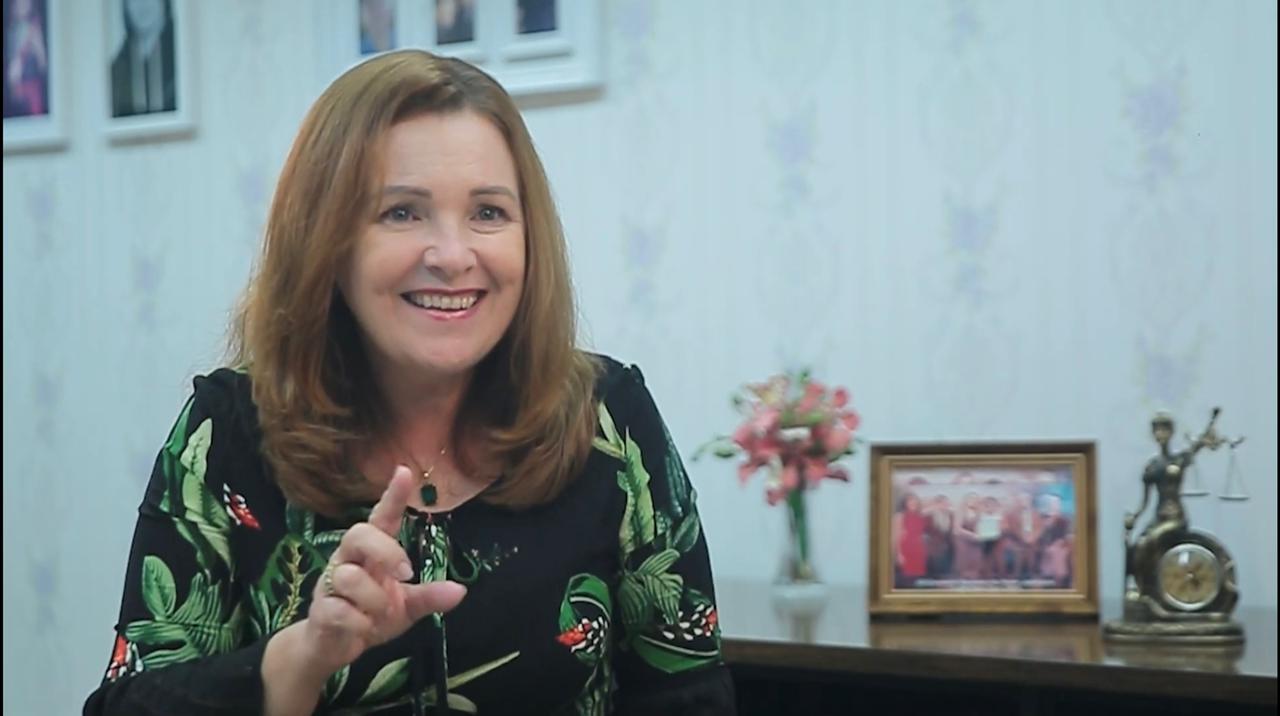 Foto retirada de vídeo da desembargadora Elaine Bianchi, sentada sorrindo, ao lado dela uma mesa com um vaso de flores, um porta-retratos e uma estátua da justiça. Na parede ao fundo aparecem parte de quadros.