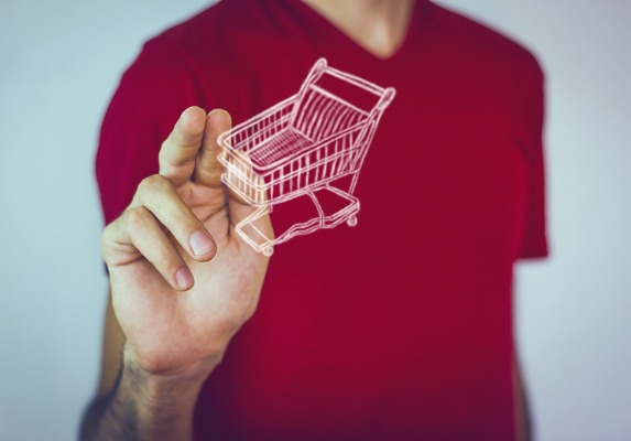 Foto do braço de um homem com a mão próxima de tocar uma ilustração de um carrinho de supermercado que está na frente dele.