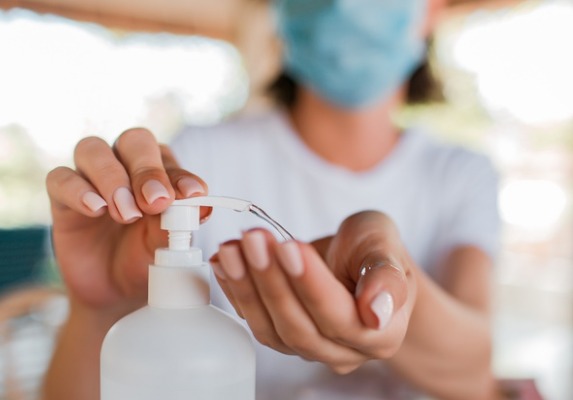 Foto de Mulher de máscara, tocando com uma mão em recipiente de álcool em gel, e colocando na outra mão.