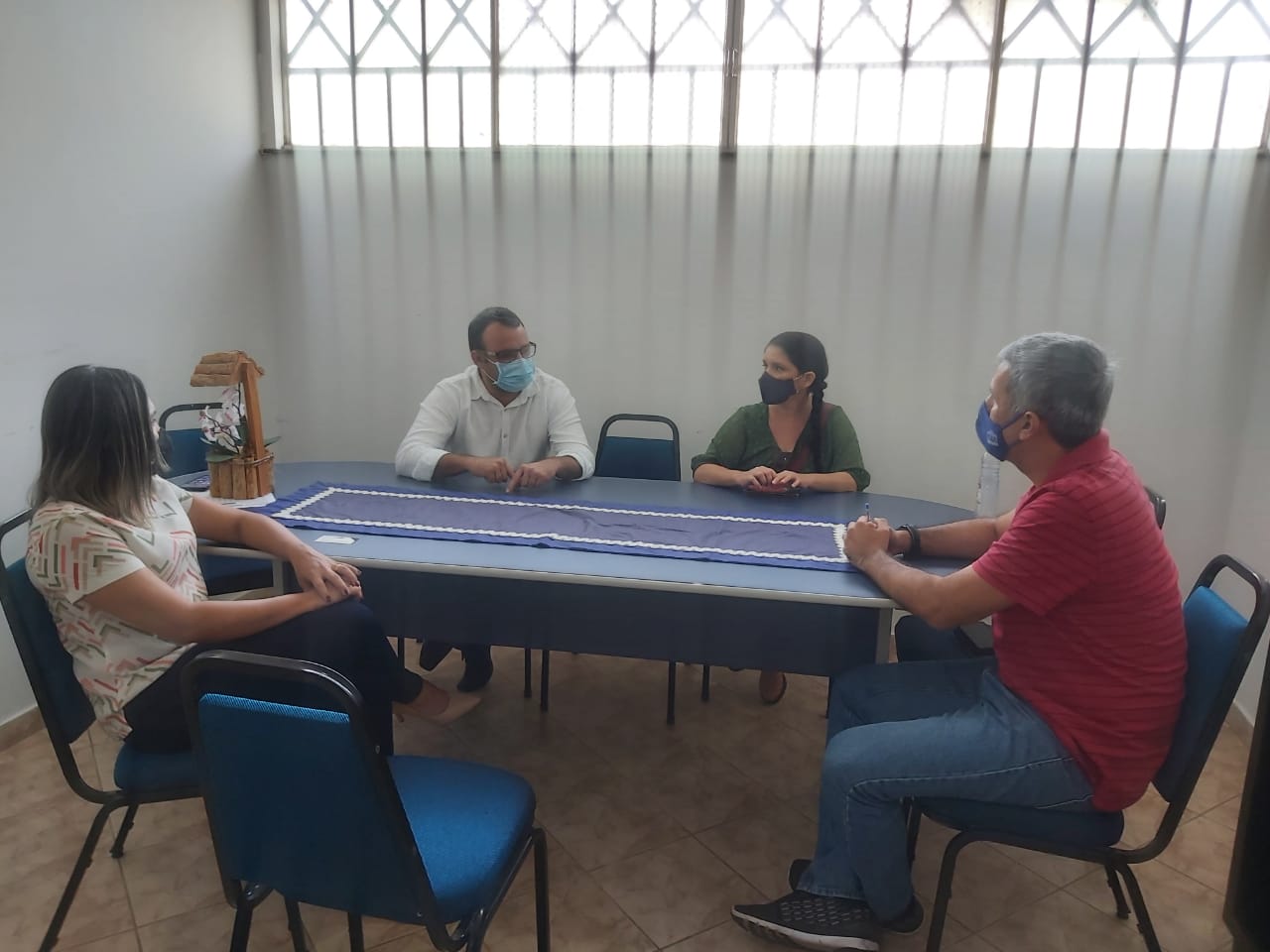 Foto de quatro pessoas sentadas em uma sala, em volta de uma mesa, conversando, em cima da mesa há uma decoração com flores e um pano.