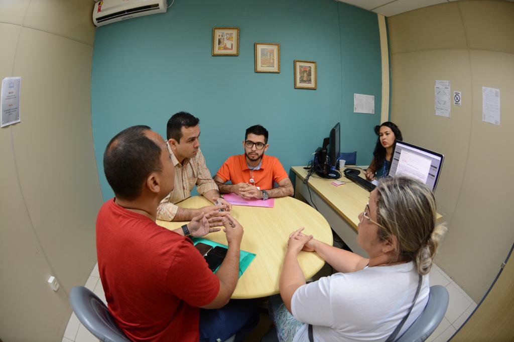 Quatro pessoas sentadas ao redor de uma mesa redonda e uma sentada atrás de uma mesa, com dois computadores em cima, no canto da sala. 