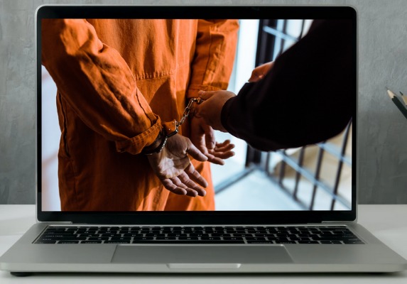 Foto de um notebook que mostra na tela a imagem de um detento de costas, vestido com as roupas da prisão e algemado e com as mãos de um policial segurando as algemas.