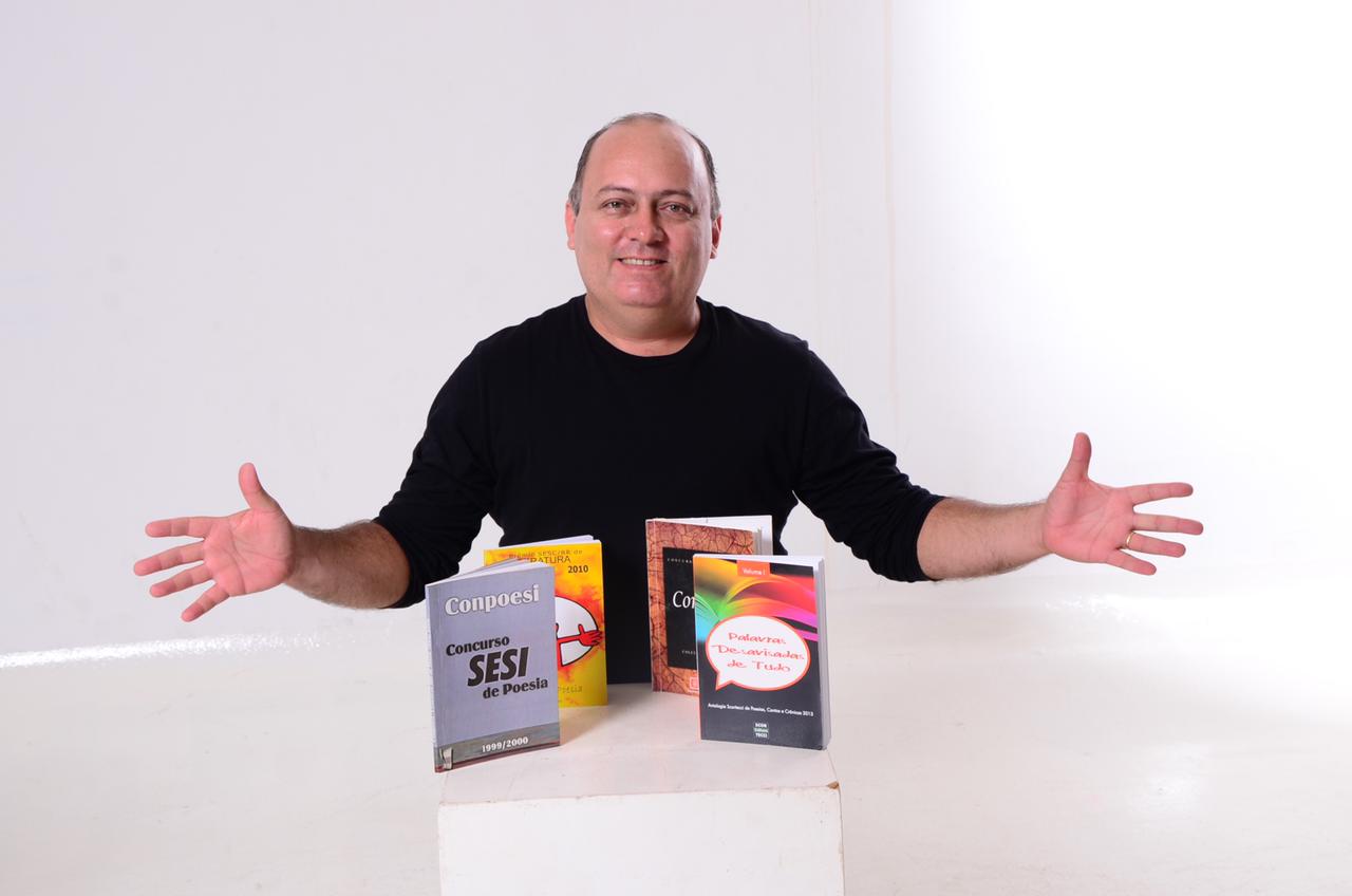 Foto de Aldair Ribeiro dos Santos, em sala branca, atrás de bancada com quatro livros de pé em cima dela.