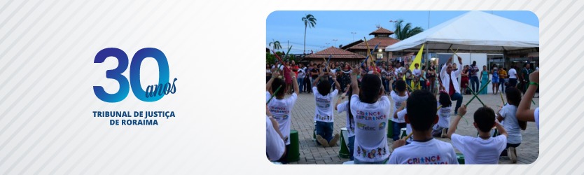 Crianças se apresentando tocando tambor em praça, com várias pessoas de pé assistindo e uma tenda branca ao fundo. e imagem dos 30 anos do TJ