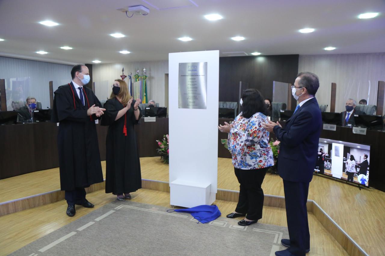 Presidente do Tribunal de Justiça e servidores de pé aplaudindo o descerramento da placa do Palácio da Justiça, que está no centro da imagem. ao fundo estão as bancadas dos magistrados.