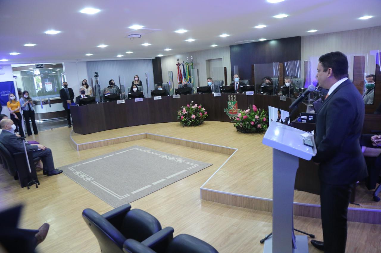 Foto de sessão no Tribunal de Justiça, com servidor falando em púlpito, magistrados sentados atrás das bancadas e cadeiras da platéia com algumas pessoas, também algumas pessoas de pé perto da porta. 