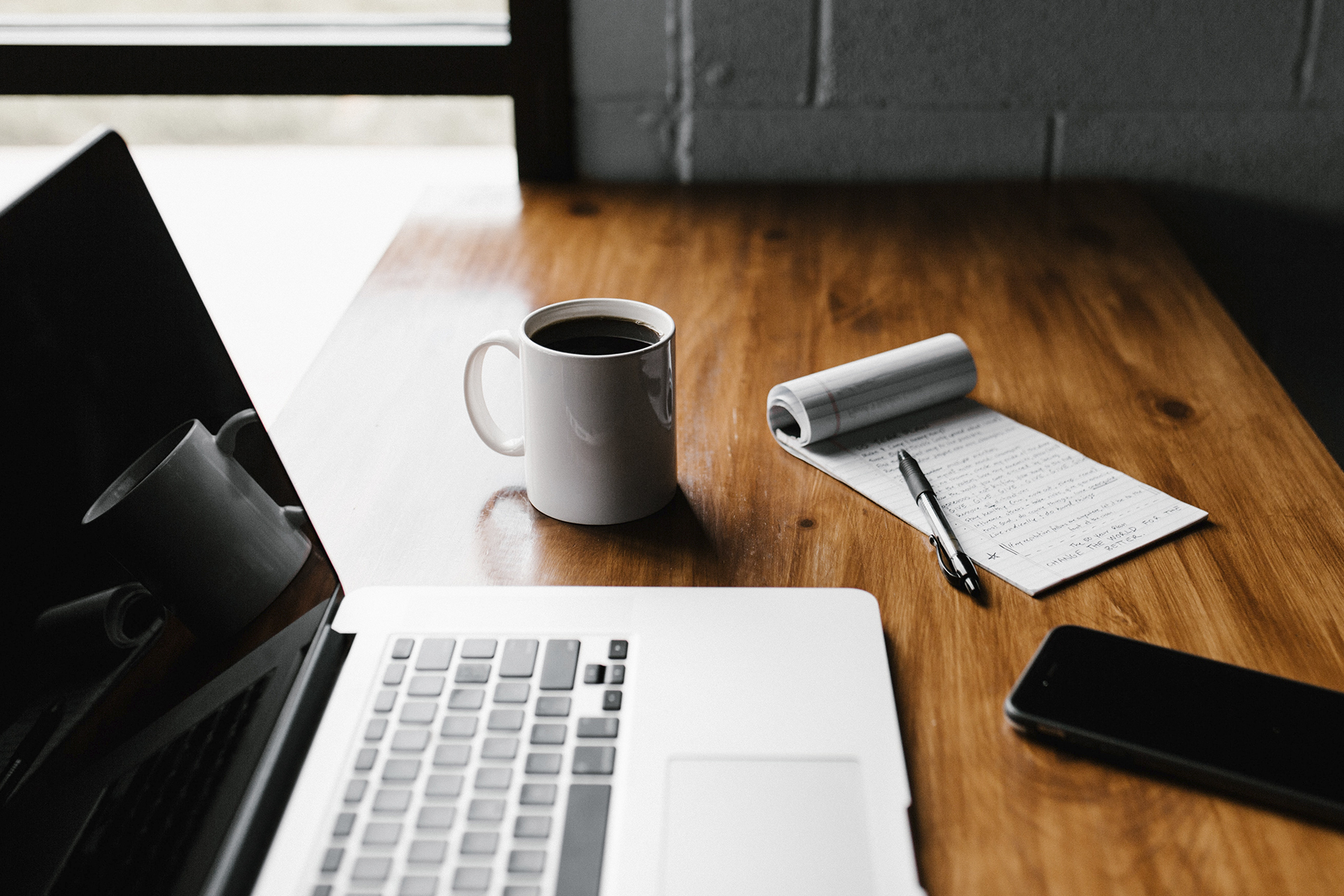 Foto de uma mesa, com um notebook, um celular, uma xícara com café, em bloco de anotações e uma caneta.