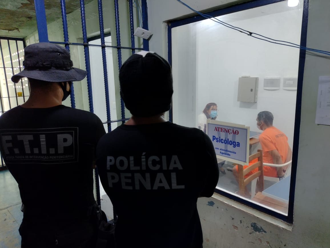 Dois policiais penais em pé de costas, olhando um reeducando tendo atendimento através de um vidro, com uma placa colada escrito “Atenção Psicóloga". o reeducando e a psicóloga estão sentados e o reeducando está com as mãos algemadas para trás.