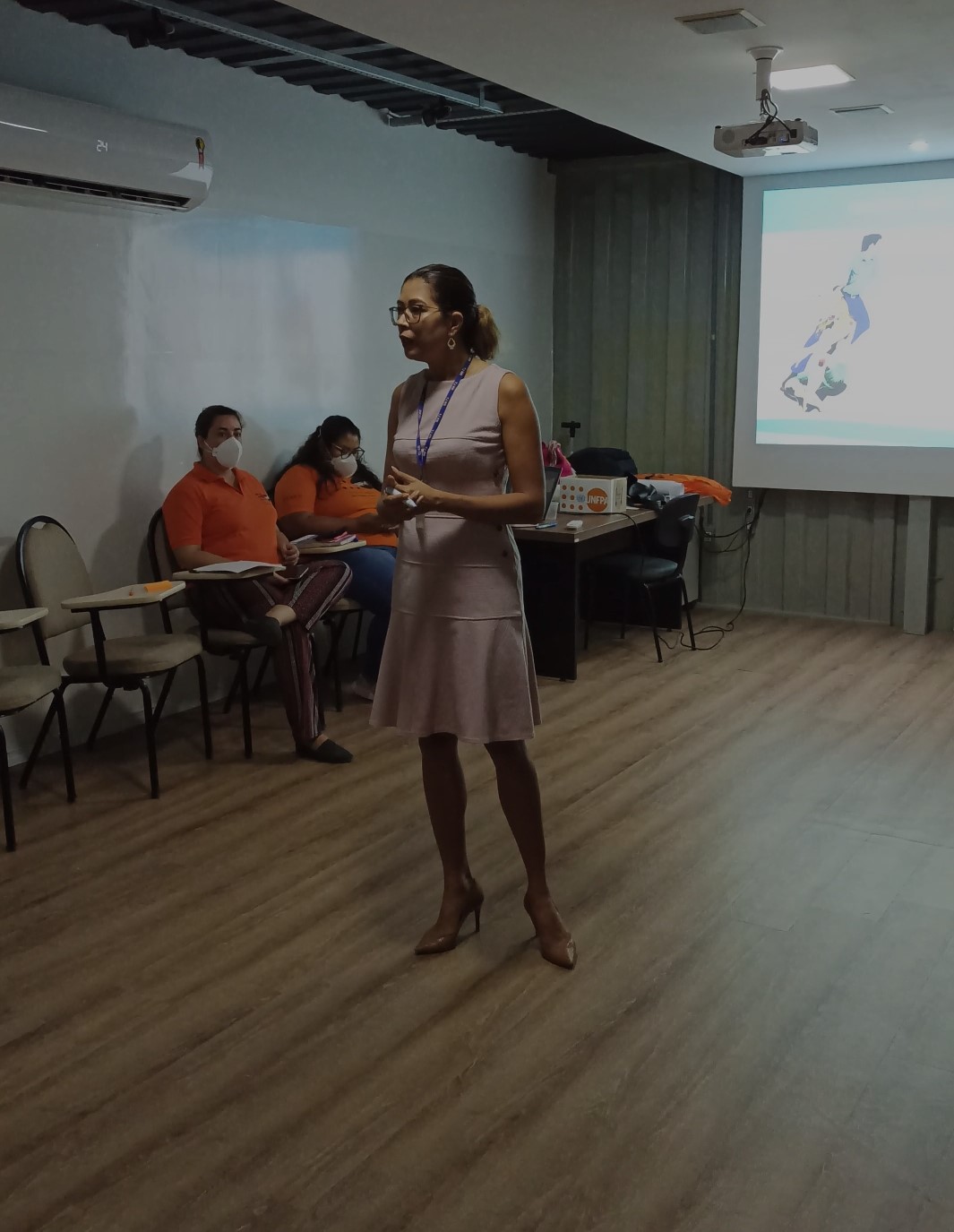 Aurilene Mesquita de pé na palestra, com servidoras sentadas em cadeiras encostadas na parede, com mesa e imagem projetada atrás dela.