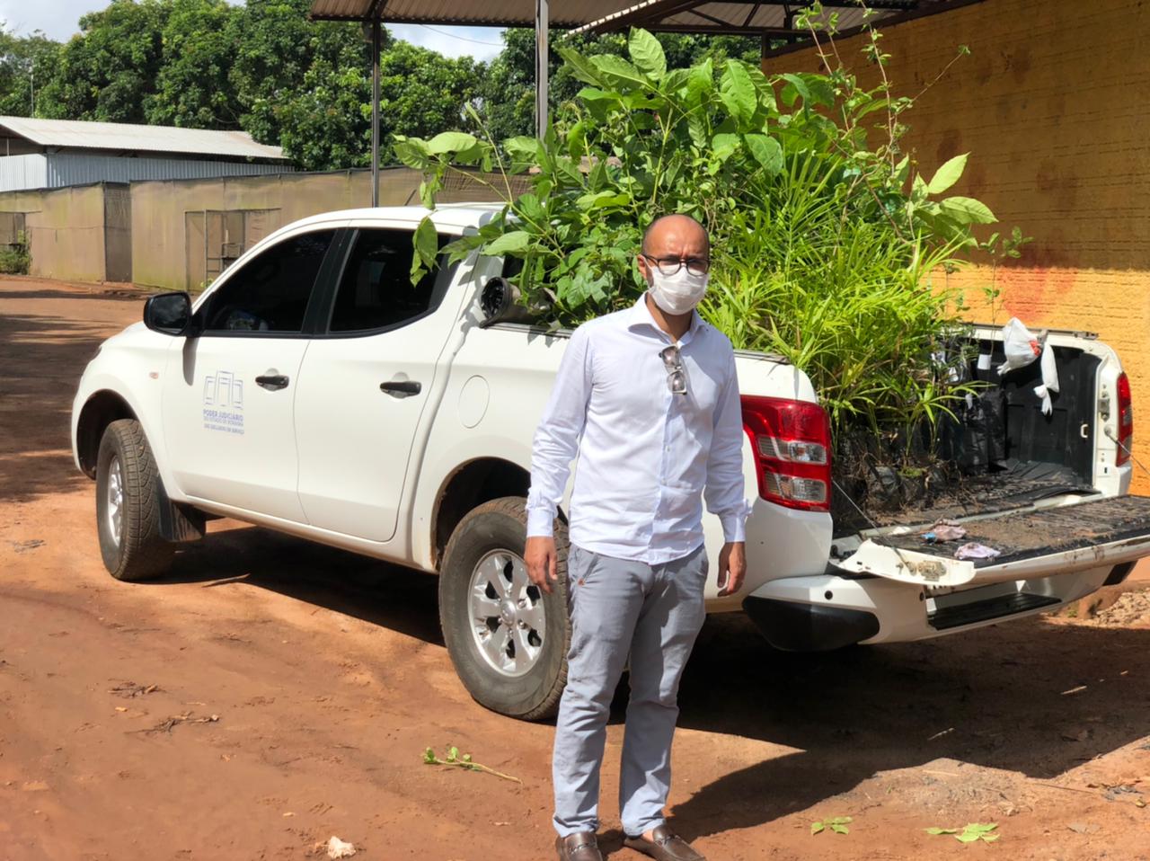 Homem de pé em frente a um carrro do Tribunal de Justiça que está com mudas de plantas dentro dele.