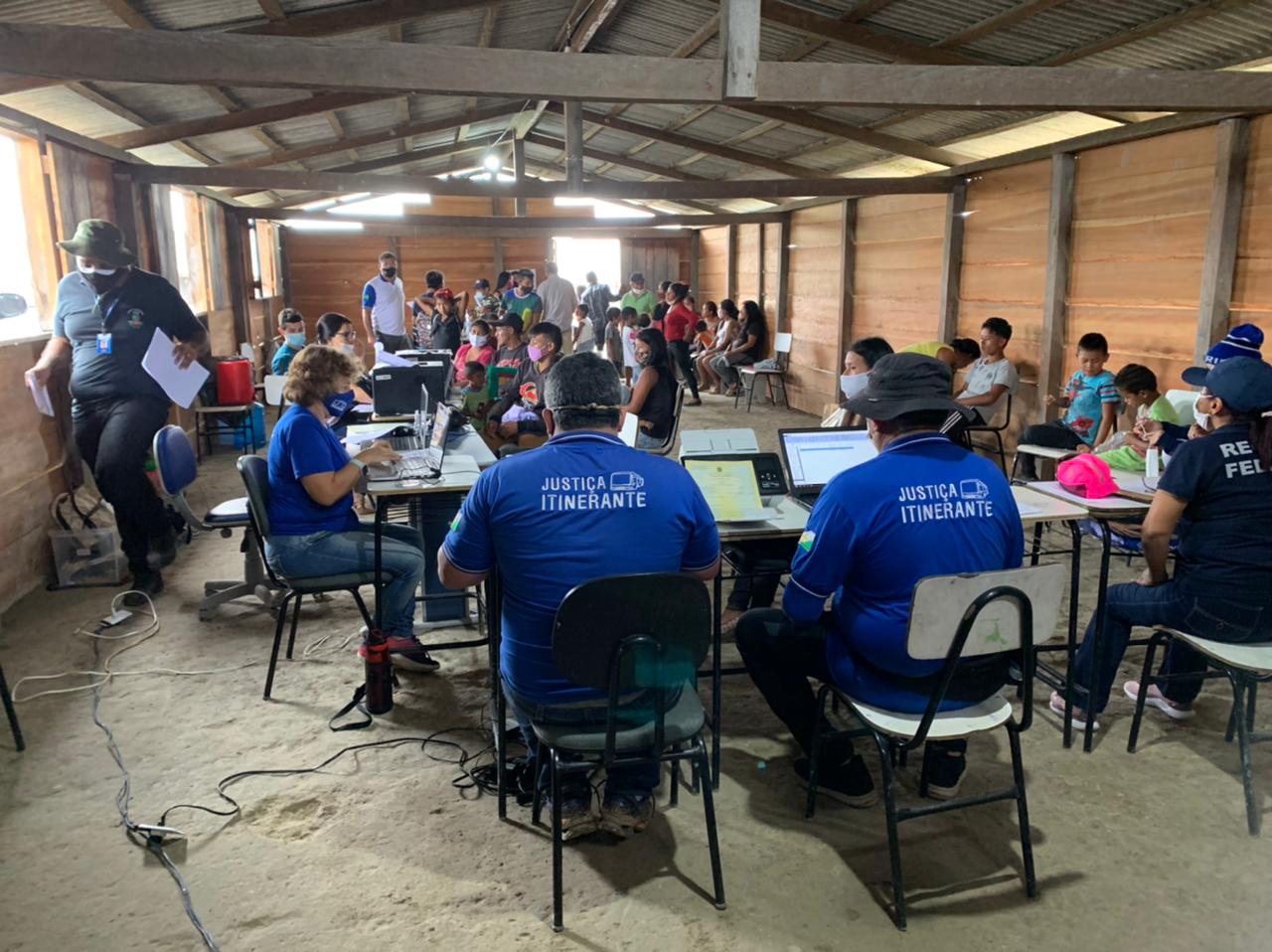 Servidores da Justiça itinerante sentados em cadeiras, com notebooks em mesas atendendo pessoas dentro de barracão e várias pessoas ao fundo esperando pelos serviços.  