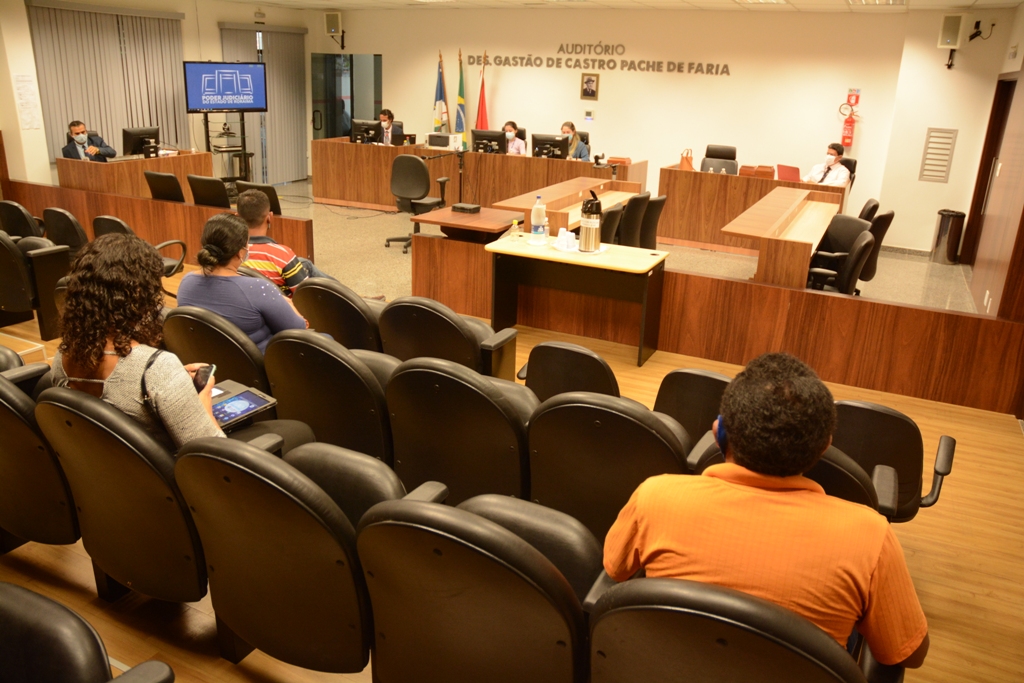 Pessoas sentadas em cadeiras assistindo sessão no auditório Gastão de Castro Pache de Faria, com a bancada dos juízes, sentados atrás de computadores ao fundo.