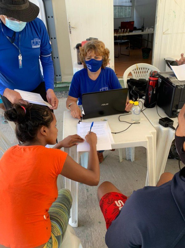 Servidores da Justiça Itinerante, um de pé segurando um papel, e a outra sentada atrás de uma mesa com um notebook e uma impressora em cima, atendendo uma mulher que está abaixada assinando um papel.