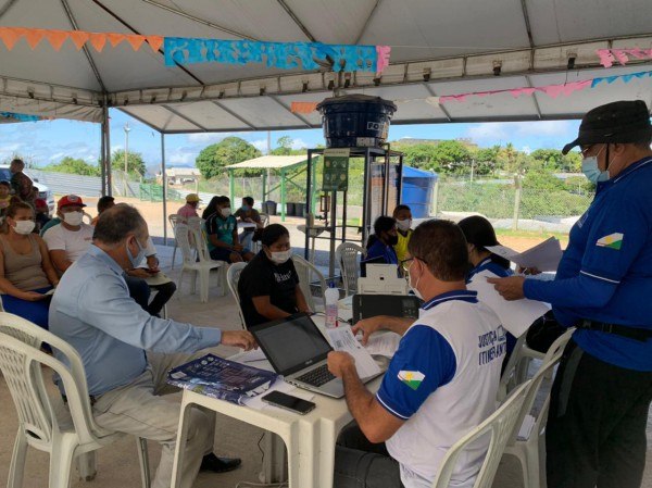 Servidores da Justiça Itinerante sentados em cadeira de plástico, com mesas de plástico com notebooks em cima, atendendo pessoas sentadas em cadeiras de plástico e outras pessoas atrás sentadas, esperando o atendimento.