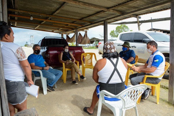 Servidores da Justiça Itinerante conversando com indígenas em frente a uma casa, sentados em cadeiras de plástico, com dois carros ao fundo.  