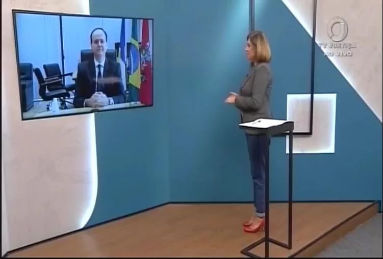 Apresentadora de TV em pé em programa, entrevistando o desembargador Cristóvão Suter, que está em uma tela participando por meio de videochamada.