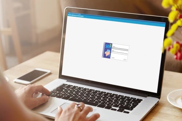 Foto de mãos de uma pessoa com uma mesa em frente, com um celular e um notebook acessando Sistema Eletrônico de Informações (SEI).