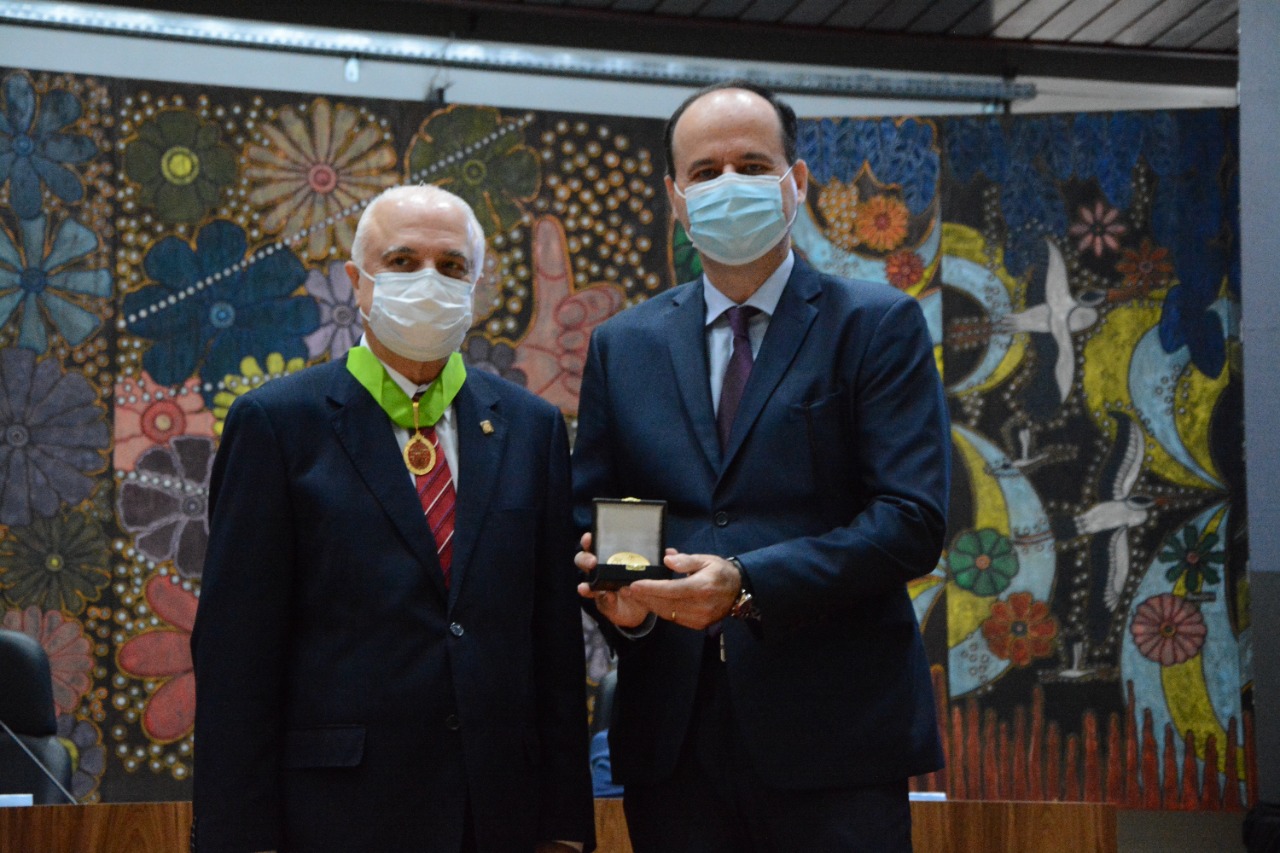 Foto colorida mostra o presidente do TJRR, desembargador Cristóvão Suter, ao lado do ministro Péricles Aurélio Lima de Queiroz, segurando o medalhão do Bicentenário do Superior Tribunal Militar.