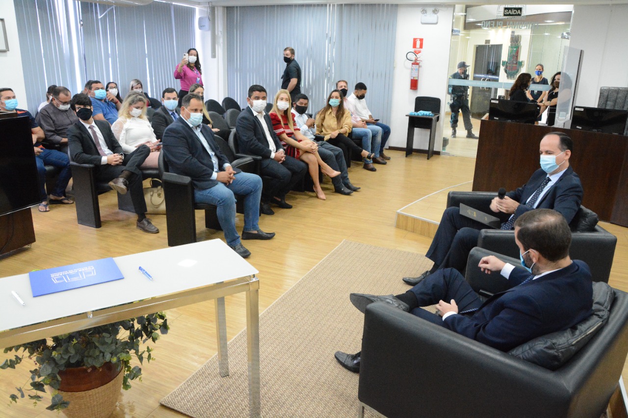 imagem colorida mostra o juiz auxiliar da presidência e o presidente do TJRR sentados em frente ao palco do auditório. À esquerda, sentados, estão prefeitos, procuradores, representantes de instituições e de municípios de Roraima. À frente está uma mesa com um vaso de plantas embaixo