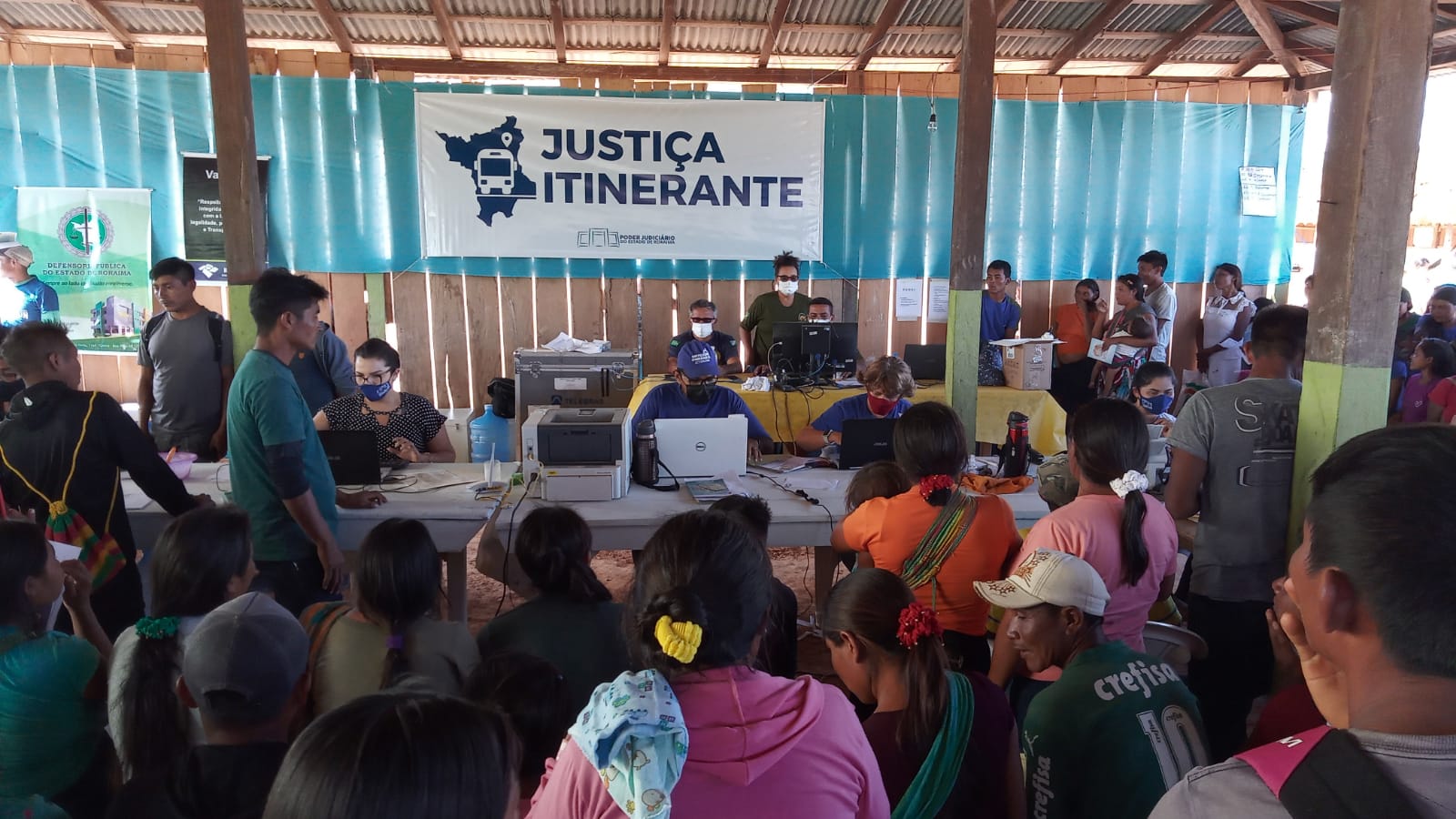 imagem colorida mostra indígenas ingaricó aguardando atendimento por parte dos servidores da Justiça Itinerante, que estão sentados frente a computadores em baixo de um barracão. Na parede do barracão há uma placa escrito “Justiça Itinerante” e “Poder Judiciário de Roraima”.