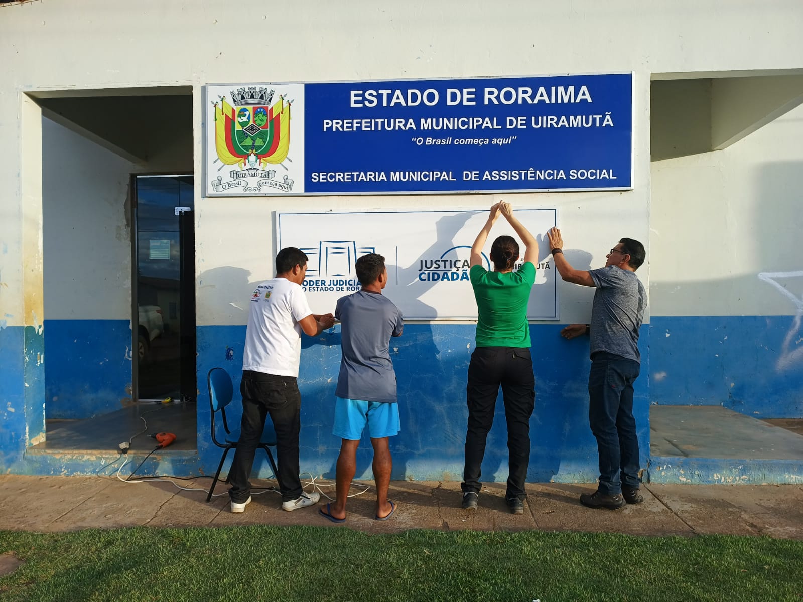  Imagem colorida contém quatro pessoas colocando a fachada do Posto Avançado de Atendimento do Programa  “Justiça Cidadã” no município de uiramutã, durante inauguração do posto.