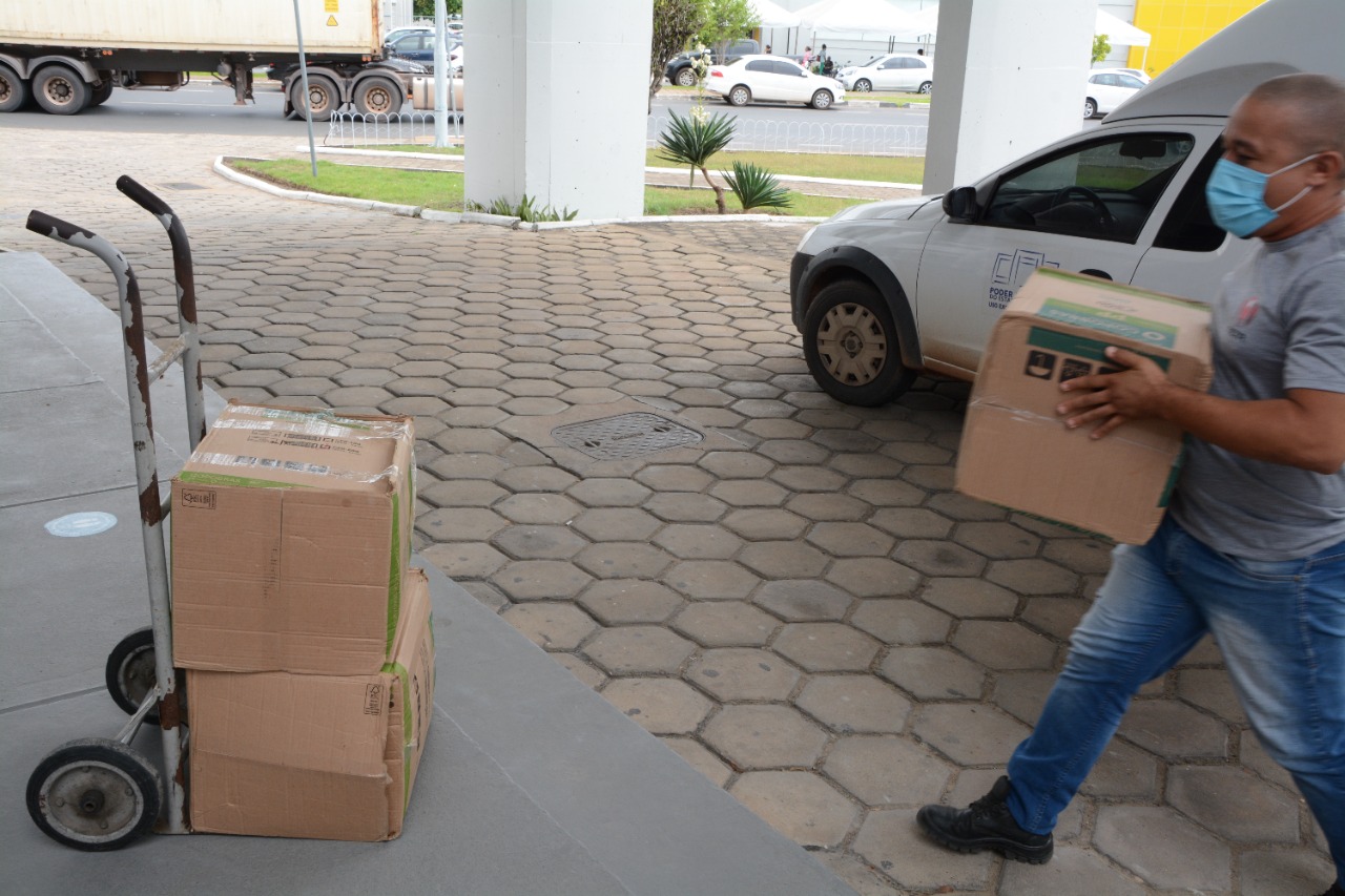 Imagem colorida mostra servidor terceirizado do TJRR em frente à entrada do Fórum Sobral Pinto, carregando caixa para colocar no carrinho de puxar peso no qual já contém mais duas caixas uma em cima da outra. 