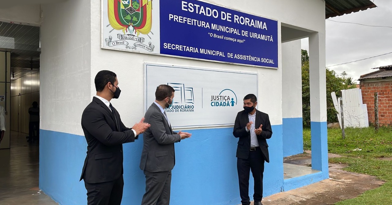 imagem colorida mostra juiz titular da comarca de Pacaraima, juiz auxiliar da presidente do TJRR e prefeito de Uiramutã. Todos estão de pé em frente a placa do Posto Avançado de Atendimento do TJRR.