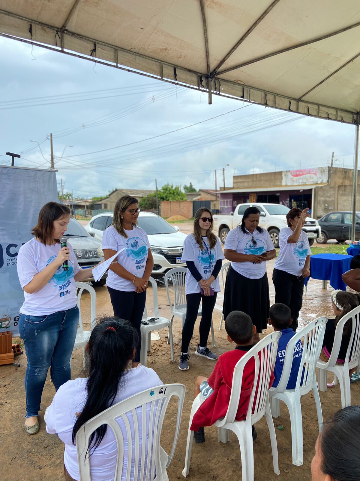 Foto colorida mostra uma mulher em pé de camisa branca e calça jeans falando ao microfone. Outras quatro mulheres de camisa branca em pé estão ao lado. Três crianças e uma mulher estão sentadas de frente para as cinco voluntárias do projeto