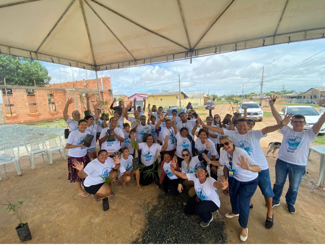 Foto colorida mostra cerca de 30 voluntários, todos de camisa branca, do projeto Bairro Sustentável embaixo de uma tenda branca, posando para foto.Alguns voluntários seguram mudas de planta nas mãos. Imagem também mostra aos fundos carros, casas, ruas sem asfalto e postes de energia.