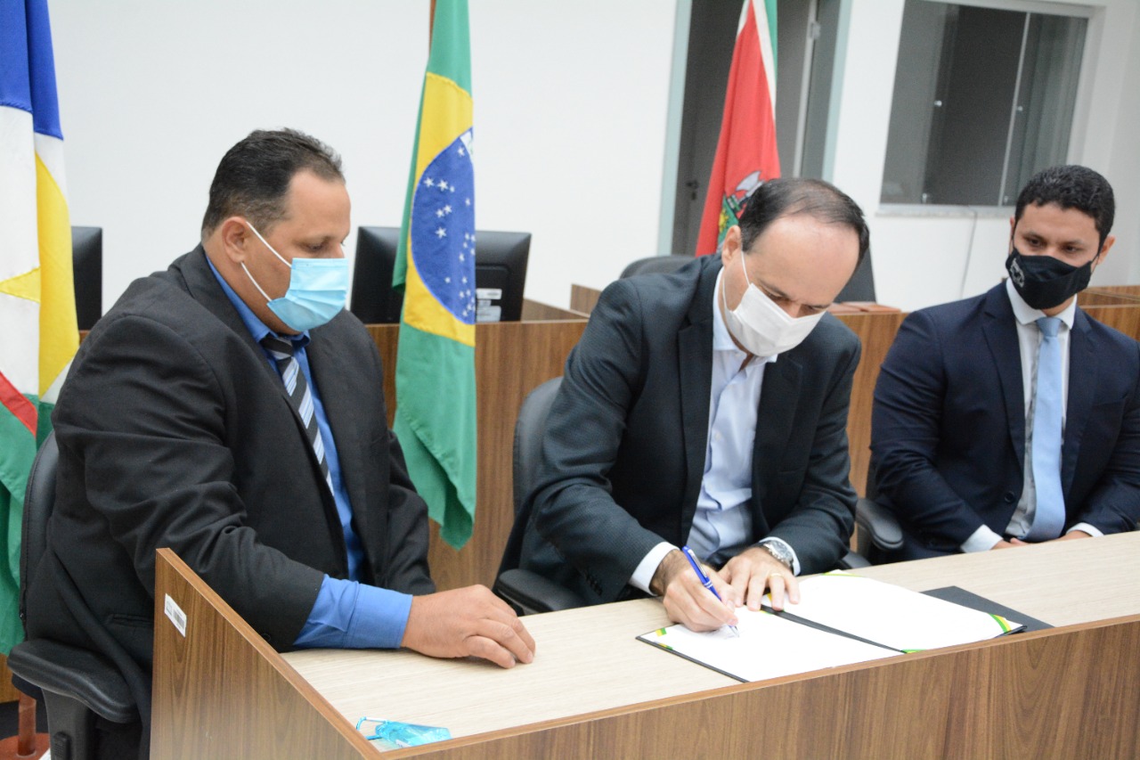 imagem colorida mostra o presidente do TJRR, desembargador Cristóvão Suter, sentado enquanto assina o termo de cooperação técnica. À direita, está o juiz titular da comarca de Pacaraima e à esquerda o prefeito de Pacaraima.